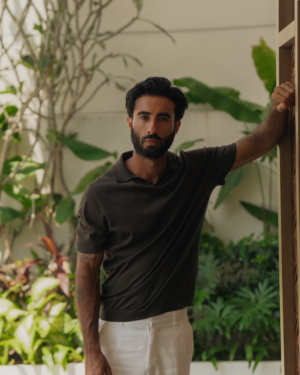Suroesh Jazayeri in a dark brown polo, leaning against a wooden frame in front of tropical plants.